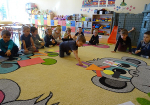 Chłopiec układa pajaca z figur geometrycznych według wzoru, dzieci siedzą wokół niego i obserwują z zainteresowaniem.